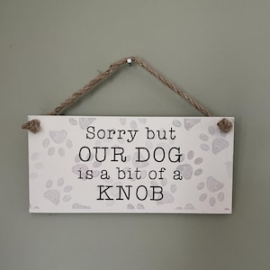 May include: A white wooden sign with a brown rope hanger. The sign has a paw print pattern and says "Sorry but OUR DOG is a bit of a KNOB".