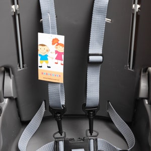 Pode incluir: Um assento infantil de plástico cinza com um sistema de cinto de cinco pontos com tiras de nylon cinza ajustáveis e uma fivela de plástico preta. Uma etiqueta branca com uma ilustração de duas crianças e o texto BABYCOVER www.babycover.cz está anexada.