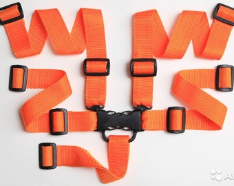 Puede incluir: Un arnés de seguridad de color naranja brillante con múltiples correas de nailon tejido y hebillas ajustables de plástico negro. El componente central es una hebilla de plástico negro con cierre lateral y anilla en D, conectada a varias correas largas.