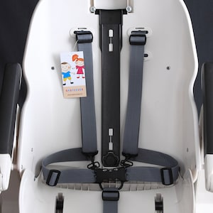 May include: A white high chair with gray straps and a black buckle. The chair has a tag with cartoon children and the text "BABYCOVER". The chair is designed for infant safety.