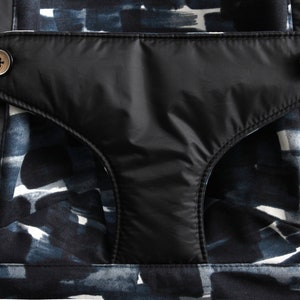 May include: Close-up of a black pocket with two wooden buttons and a gray strap detail. The pocket is set against a fabric with a blue and white abstract pattern. The overall design is modern and stylish.