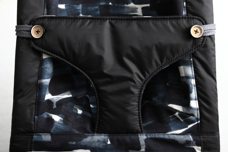 May include: Close-up of a black pocket with two wooden buttons and a gray strap detail. The pocket is set against a fabric with a blue and white abstract pattern. The overall design is modern and stylish.