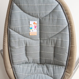 Pode incluir: Uma capa para assento de baloiço de bebé em tecido cinzento com riscas brancas e uma borda bege. O assento tem um design acolchoado e uma etiqueta com ilustrações de desenhos animados e o texto "BABYCOVER".