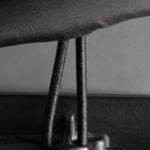 May include: Close-up of a black fabric with a textured surface, featuring two black elastic cords and a plastic clip. The cords are attached to the fabric, suggesting a fastening mechanism or closure system.