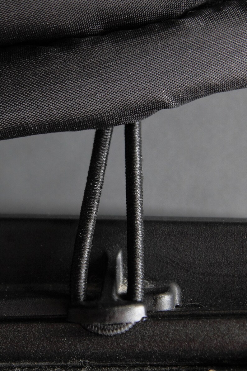May include: Close-up of a black fabric with a textured surface, featuring two black elastic cords and a plastic clip. The cords are attached to the fabric, suggesting a fastening mechanism or closure system.