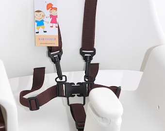 Puede incluir: Un arnés de seguridad marrón de cinco puntos con hebillas de plástico negro y correas ajustables fijado a una trona blanca. Una etiqueta rectangular está sujeta a la correa izquierda con ilustraciones y el texto BABYCOVER.