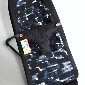 May include: A black baby bouncer with a patterned fabric insert featuring a design of dark blue and white brushstrokes. The bouncer has a black frame and base, with a tag attached. The bouncer is designed for infants.