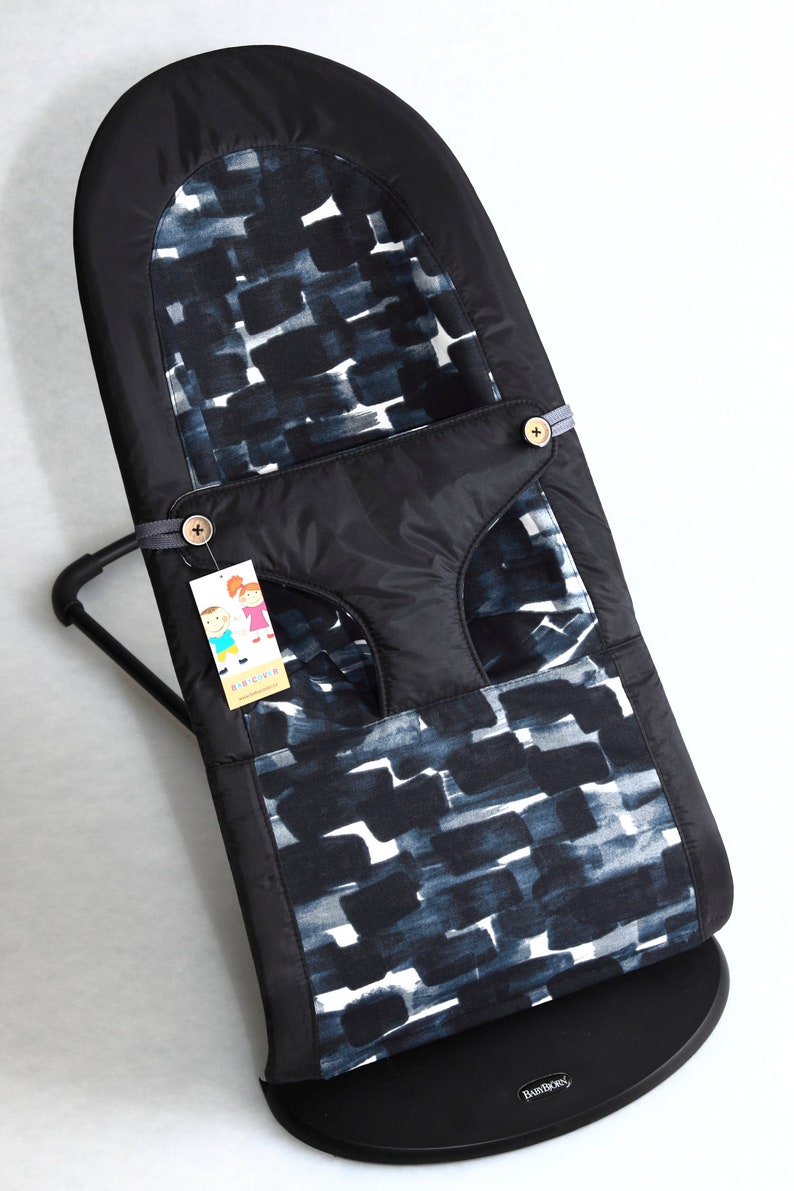 May include: A black baby bouncer with a patterned fabric insert featuring a design of dark blue and white brushstrokes. The bouncer has a black frame and base, with a tag attached. The bouncer is designed for infants.