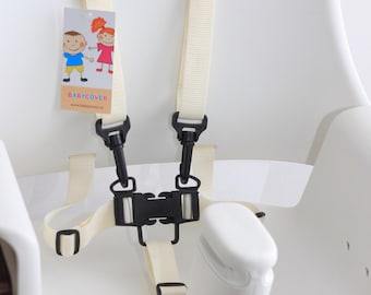 Puede incluir: Un arnés de seguridad blanco para trona con correas de nailon color crema, hebillas de plástico negro y un cierre central. Se adjunta una etiqueta rectangular con una ilustración de dos niños y el texto BABYCOVER.