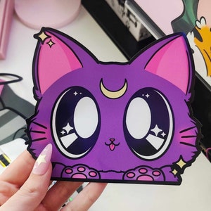 May include: A purple cat with big eyes and a crescent moon on its forehead. The cat has pink ears and paws and is surrounded by sparkling stars.
