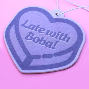 May include: A heart-shaped air freshener in shades of purple, with the text "Late with Boba!" in white. The air freshener is suspended by a white string against a pink background. The design features a heart shape with a border.