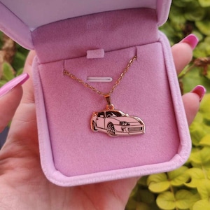 May include: A gold-colored necklace with a car pendant in a pink velvet box. The car pendant is a white silhouette of a sports car.