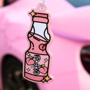 May include: A pink air freshener shaped like a Japanese soda bottle with peach illustrations and Japanese text. The air freshener is hanging by a string, with a pink car in the background.