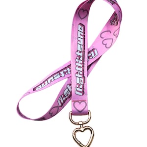 Op de afbeelding: Roze lanyard met een hartvormige sluiting. De lanyard heeft de tekst "night:tsune" erop gedrukt in een wit, gestileerd lettertype. De lanyard is versierd met kleine roze hartjes.