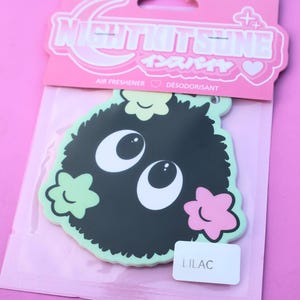 May include: A black air freshener with a cartoonish design, featuring large white eyes and a fluffy body, accented with light green and pink floral shapes. The packaging reads "NIGHTKITSUNE" and "LILAC".