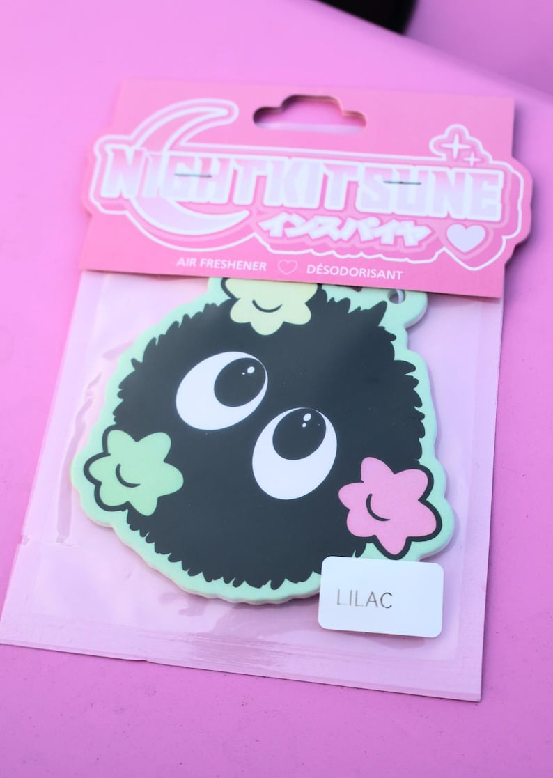 May include: A black air freshener with a cartoonish design, featuring large white eyes and a fluffy body, accented with light green and pink floral shapes. The packaging reads "NIGHTKITSUNE" and "LILAC".