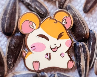 Puede incluir: Un pin de esmalte dorado con un hámster de dibujos animados con un cuerpo amarillo y blanco, mejillas rosadas y una nariz negra. El hámster está sosteniendo una semilla de girasol en sus patas y tiene una expresión feliz en su rostro.