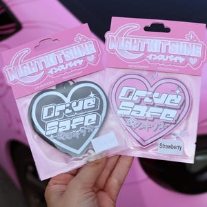 May include: Two heart-shaped air fresheners in pink packaging labeled NIGHTKITSUNE. One is black with white text reading Drive Safe and Japanese characters, the other is pink with white text reading Drive Safe and Japanese characters, labeled Strawberry.