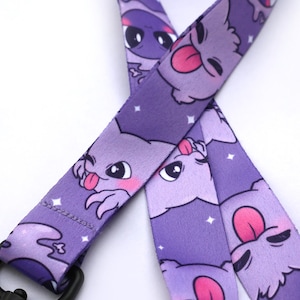 May include: Purple lanyard with a repeating pattern of cartoon purple creatures with pink tongues and white stars. The lanyard has a black metal clip.