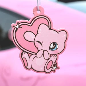 Puede incluir: Ambientador rosa con un personaje de dibujos animados con un diseño en forma de corazón. El personaje guiña un ojo azul y tiene una estrella en la mejilla. Colgando de un cordón blanco.