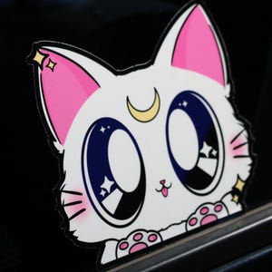 May include: A white cartoon cat with pink ears and a crescent moon on its forehead. The cat has large blue eyes with white stars inside. The cat is looking at the viewer and has a pink tongue sticking out. The cat is on a black background.
