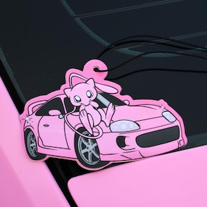 May include: A pink car air freshener depicting a cartoon character on a pink sports car. It has a black string for hanging. The background shows a car's windscreen and bonnet.