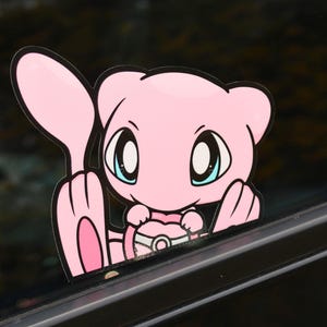 Puede incluir: Personaje de dibujos animados rosa con ojos grandes y una Poké Ball rosa. El personaje está mirando desde detrás de una superficie negra.