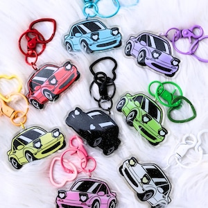 May include: A set of nine keychains featuring colourful cartoon cars with heart-shaped headlights. Each keychain has a different colour car and a different style of metal ring and clasp.