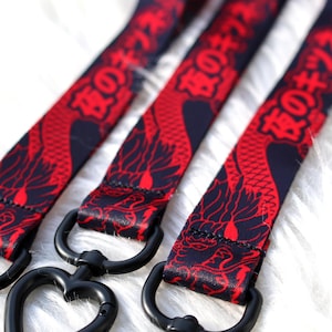 Pode incluir: Close-up de três cordões com um design de dragão vermelho sobre um fundo azul marinho. Cada cordão tem um fecho preto em forma de coração. Os cordões estão sobre uma superfície branca e fofa.