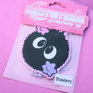 May include: A pink air freshener package featuring a black cartoon character with large white eyes and a purple flower. The package has the text "NIGHTKITSUNE" and "Strawberry".