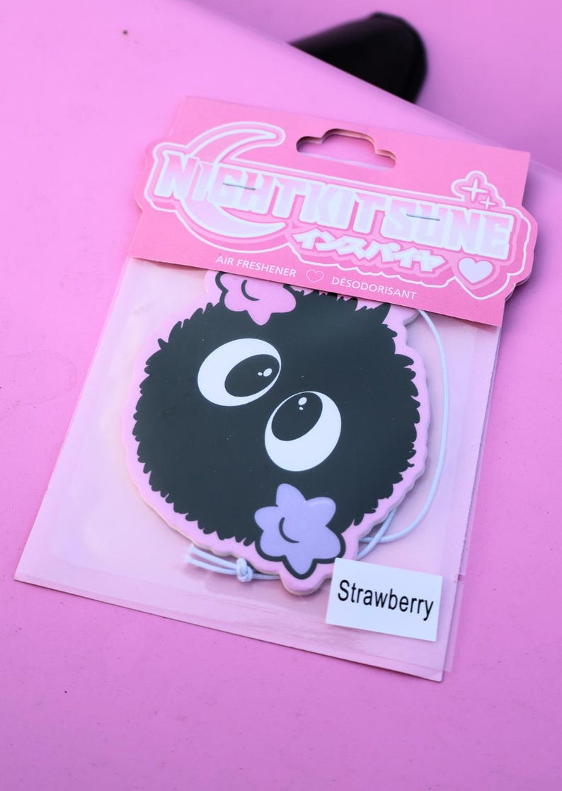 May include: A pink air freshener package featuring a black cartoon character with large white eyes and a purple flower. The package has the text "NIGHTKITSUNE" and "Strawberry".