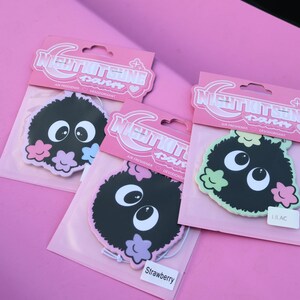May include: Three air fresheners with a cartoon character design. Each air freshener has a black, fuzzy body, large white eyes, and colorful flower accents. The packaging is pink with Japanese text.