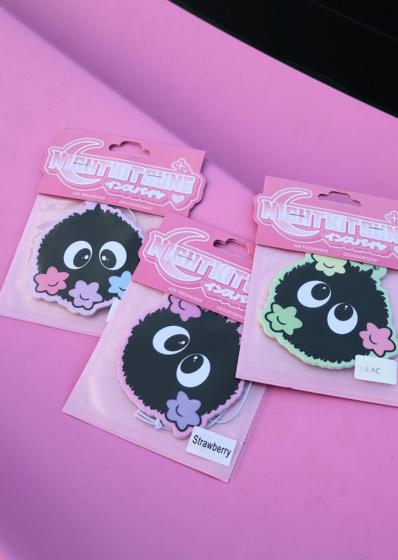May include: Three air fresheners with a cartoon character design. Each air freshener has a black, fuzzy body, large white eyes, and colorful flower accents. The packaging is pink with Japanese text.