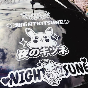 Puede incluir: Un coche negro con una pegatina blanca en la ventana trasera. La pegatina presenta un personaje de dibujos animados con el texto "Night Kitsune" y caracteres japoneses.