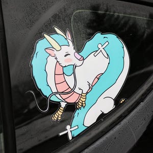 May include: A blue and white cartoon dragon sticker on a black car window. The dragon is curled up with its tail wrapped around its body. The sticker has a pink and white design.