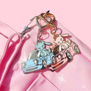 May include: Two metal keychains featuring a cartoon character in a car. One keychain is light blue with a blue car, and the other is pink with a pink car. Both have silver and gold-colored clasps.