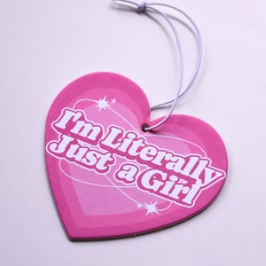 May include: A pink heart-shaped tag with the text "I'm Literally Just a Girl" in white. The tag has a light pink gradient and a white string for hanging. Decorative stars and lines are also visible.