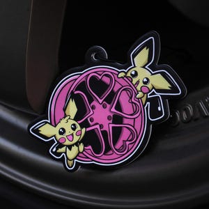 May include: A black and pink charm featuring two yellow cartoon characters with black outlines. The charm has a pink wheel design with heart shapes. The charm is set against a dark background.