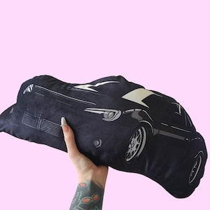 May include: A black and white car pillow. The pillow is made of a soft, plush fabric and features a detailed illustration of a car. The pillow is perfect for adding a touch of style to any room.