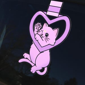 May include: A purple cat with a crescent moon on its forehead is trapped inside a heart-shaped frame. The frame is attached to a purple and white striped ribbon.