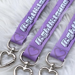 Op de afbeelding: Paarse lanyards met de tekst "Nightkitsune" in het wit, met een hartafbeelding. Elke lanyard heeft een zilverkleurige hartvormige sluiting. De lanyards worden weergegeven op een zacht, wit oppervlak.