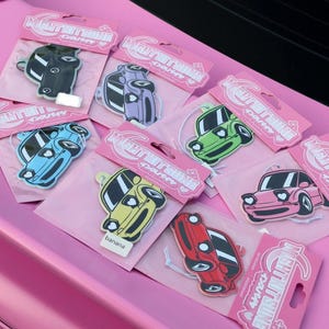 Puede incluir: Una colección de ambientadores con forma de coche en varios colores, incluyendo negro, morado, verde, azul, amarillo, rosa y rojo. Cada ambientador está empaquetado en una funda de cartón rosa etiquetada Night Kitsune con un cordón blanco.