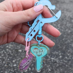 Op de afbeelding: Een lichtblauwe sleutelhanger met een sleutelvormige bedel met de tekst "DRIVE SAFE" in witte letters. De sleutelhanger heeft ook een roze hartvormige bedel met tekst. De sleutelhanger wordt door een persoon vastgehouden.