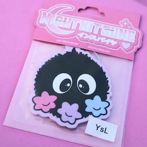 May include: A black air freshener with a cartoon-like design featuring large white eyes and three flower-shaped accents in pink, purple, and blue. The packaging includes the text "NIGHTKITSUNE" and "AIR FRESHENER". A small white label reads "YsL".