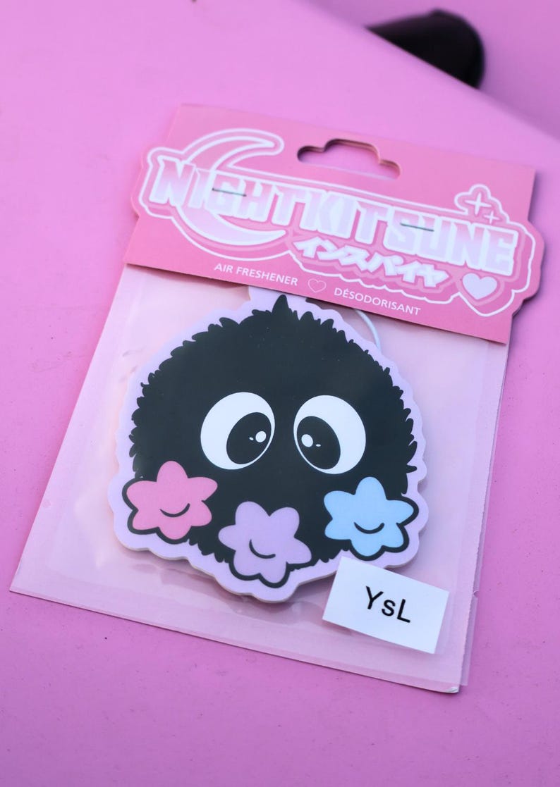 May include: A black air freshener with a cartoon-like design featuring large white eyes and three flower-shaped accents in pink, purple, and blue. The packaging includes the text "NIGHTKITSUNE" and "AIR FRESHENER". A small white label reads "YsL".