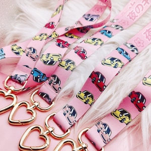 May include: Several pink lanyards featuring a repeating pattern of cartoon cars in various colours and Japanese text. Each lanyard has a gold-coloured heart-shaped clasp. The lanyards are displayed on a soft, white, furry surface.