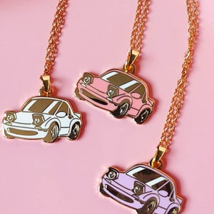 Puede incluir: Tres collares de oro con dijes de coches. Los dijes son de color rosa, blanco y morado. Los collares están sobre un fondo rosa.