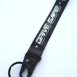 May include: Black lanyard featuring the white text "DRIVE SAFE!" and Japanese characters. It has a black metal clasp and a split ring. The background is plain white. Ideal for keys or accessories.