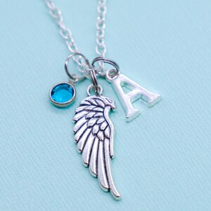 May include: Silver-toned necklace with a chain and multiple charms. Charms include a wing, a letter "A", and a small blue gemstone. The necklace is displayed against a light blue background.