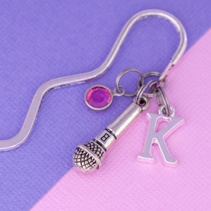 May include: Silver bookmark with a pink rhinestone, a microphone charm, and a letter K charm. The bookmark is on a purple and pink background.